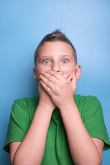 Studio shot of emotional adorable boy  covering open mouth with hand being surprised and shocked, showing true emotion. Portrait on blue background 