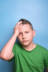 Studio shot of emotional adorable boy  covering head with hand being tired, headache,   showing true emotion. Portrait on blue background 