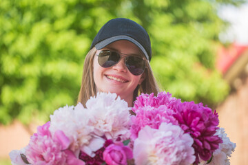 Fototapeta premium Beautiful and happy young girl in a cap and sunglasses in summer with a beautiful bouquet of peonies in nature. Close up portrait