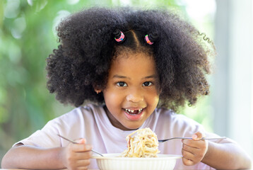 A little curly-haired African American girl sits at the table eating delicious Spaghetti Carbonara. Fun, cheerful. Appetite. Childhood and eating concepts.