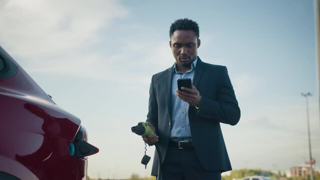 Close up of african businessman unplugging charging cable from electric car while using modern smartphone. Concept of lifestyles, ecology vehicle and busy people. Plugging in power cord to an car.