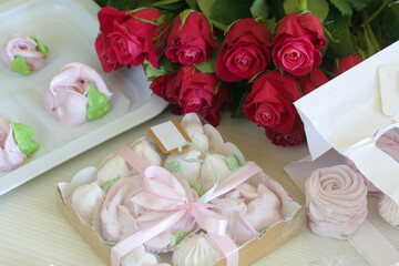 Homemade marshmallows in a gift box. Zephyr in different shapes, classic and in the form of flowers. Nearby is a bouquet of roses. Lies on a white surface.