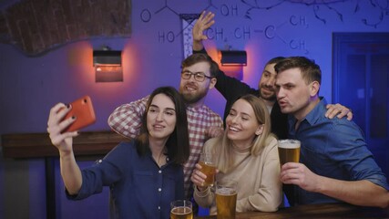 Friends in pub on bar counter with beer glasses in hands make group selfie. People watch tv and cheer for football team. Social life