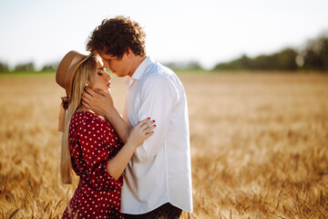 Portrait of a couple in love against the backdrop of the setting sun. A girl and a guy in wheat field. A man in white shirt and a woman in a dress with polka dots pose in the middle of a summer field.