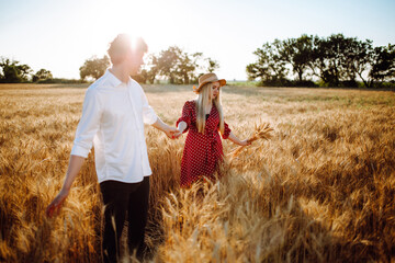 A young couple walks at sunset in a wheat field. A girl in a pea dress and a straw hat and a young man in a white shirt on blue sky background. Summer landscape.Two lovers on the wheat field.