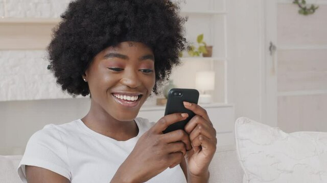 Smiling African American millennial woman holding smartphone having fun playing mobile games enjoy technology online shopping. Happy mixed race lady surfing social media mobile apps content on phone