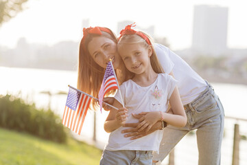 Obraz premium Patriotic holiday. Happy family, mother and daughter with American flag outdoors on sunset. USA celebrate independence day 4th of July.