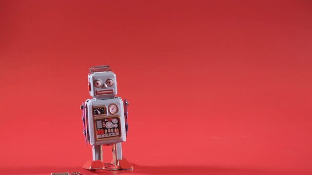 A cute tin toy robot, walking over a red backdrop, losing power and stopping (completely discharged and without the key).
