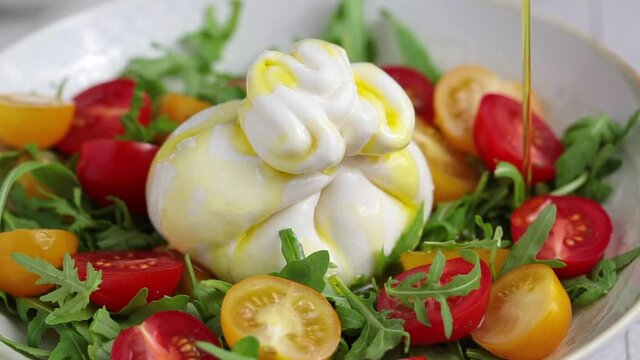 Burrata cheese salad with arugula and tomatoes pouring with olive oil, slow motion.