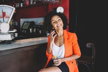 Cheerful young ethnic lady talking on smartphone while drinking cocktail in bar
