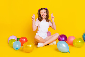 Fototapeta na wymiar Portrait of pretty cheerful girl sitting on floor with air balls decor showing v-sign isolated over bright yellow color background