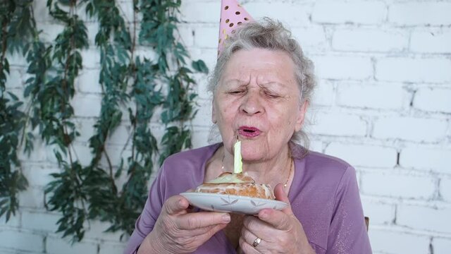 Slow motion. Happy smiling senior grandmother celebrate 85th birthday, holds cake and blowing out candle feels gratitude, white brick background. Enjoy, respect and warm relation. Celebration concept