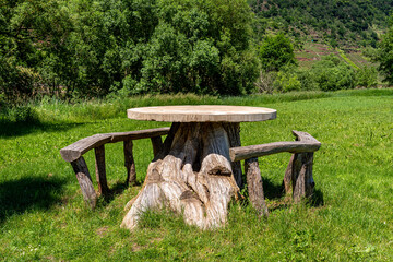 A table made of a tree trunk with a concrete top and wooden seats standing on a meadow by the river.