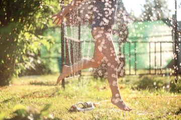 Small girl playfully jumping through sprinkler and water - obrazy, fototapety, plakaty