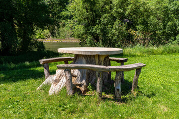 A table made of a tree trunk with a concrete top and wooden seats standing on a meadow by the river.