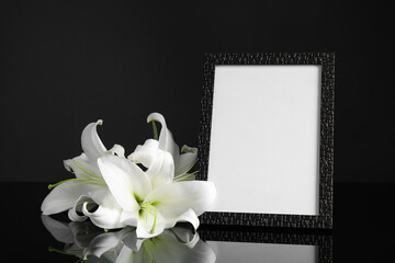 Funeral photo frame and white lilies on black table against dark background. Space for design
