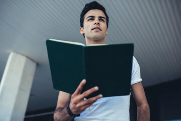 Below view of smart male student pondering on education information from textbook standing outdoors, intelligent man 20s holding notepad for learning and studying and thinking during daytime