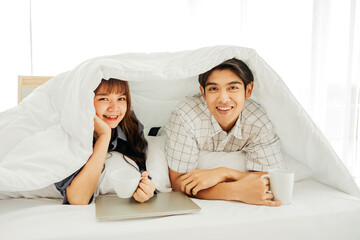 Asian young couple smiling and drinking coffee enjoy together with a laptop on the bed under white blanket at morning, lover lifestyle modern holiday concept.