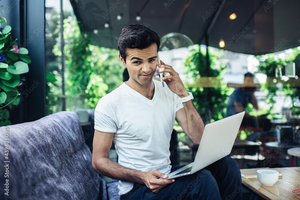 Poster portrait of millennial hipster guy with digital laptop computer looking at camera while calling via 