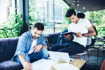 Fototapeta premium Caucasian male IT professionals analyzing paperwork during remote collaboration in street cafe, young skilled student with modern laptop technology e learning and preparing to exams together
