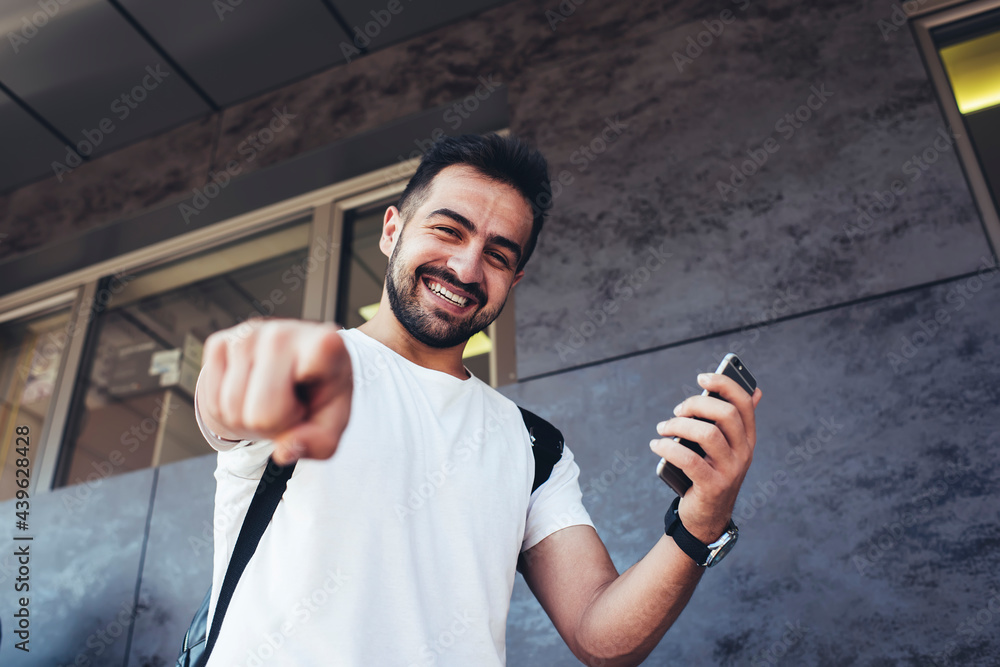 Poster half length portrait of cheerful male tourist smiling at camera while pointing at you using modern c