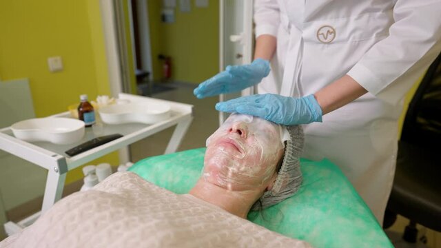 Cosmetologist applying wet disposable towels on woman face with cooling cosmetic mask layer to soothe sensitive skin after peeling, young female relaxing during professional beauty procedure in clinic