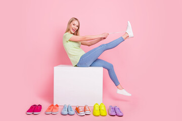 Photo of cheerful blonde lady sit podium tie shoelaces have fun wear green t-shirt jeans isolated on pink background