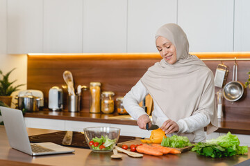 Relaxed mature arabian muslim woman in hijab cutting vegetables for salad, preparing cooking...