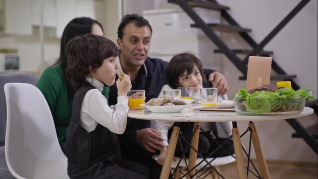 Side view of absorbed little boy eating sweet tasty bakery with blurred joyful mother father and twin brother laughing watching comedy movie on laptop at background. Happy family dinner indoors