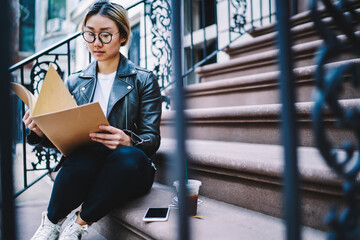 Trendy dressed female student dressed in stylish leather jacket sitting at urban stairs and checking education information noted in textbook, intelligent hipster girl in optical spectacles