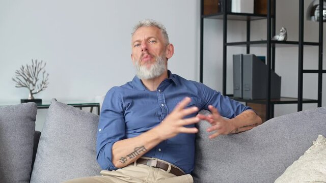 Confident senior grey haired man looks at the camera and talking, explaining something, mature male in smart casual shirt recording video, streaming online, headshot, webcam view
