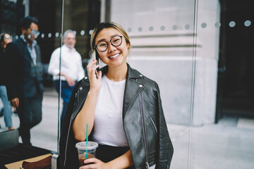 Portrait of cheerful Asian woman in optical eyewear enjoying mobile conversation while waiting for bus transport on stop bench,smiling Chinese girl talking via mobile application connecting to roaming