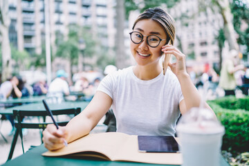 Portrait of cheerful hipster girl in spectacles smiling at camera while learning in street cafe and making international cell conversation for discussing education information, roaming connection