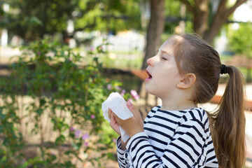 A girl with blond hair in a striped T-shirt sneezes near a bush with flowers in the park holding a napkin in her hand. Seasonal bloom allergy.