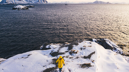 Back view from drone of female traveler in yellow winter jacket walking on snowy island cliff while looking at breathtaking nordic landscape, woman wanderlust exploring Norway fjords during journey