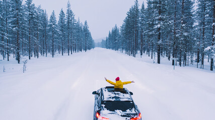 Lapland / Finland - 01.12.2020: Bird's eye view, female tourist into hatch of car raised arms enjoying winter trip in north country while driving on snowy road surrounded by coniferous forest