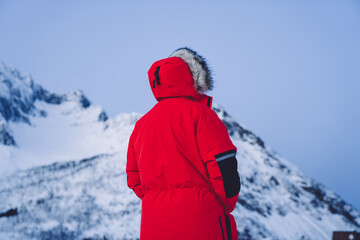 Fototapeta na wymiar Back view of male explorer in red winter coat standing near high mountains explore location on weekends trip, man traveler looking at scandinavian fjords landscape on expedition