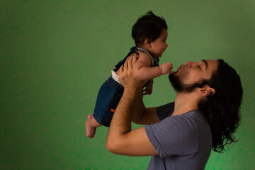 Hispanic young father with long hair and casual clothes with his newborn son hugging and happy on a green background