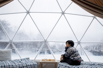 Thoughtful woman in spherical camp cupola in winter forest sitting inside of a  transparent tent with a phone 