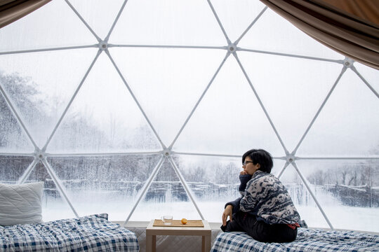 Thoughtful woman in spherical camp cupola in winter forest sitting inside of a  transparent tent with a phone 