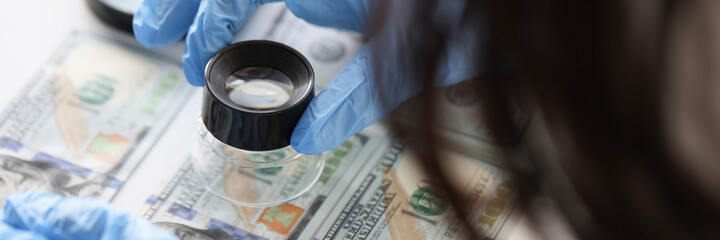 Hands in rubber gloves holding magnifying glass over dollar bills closeup