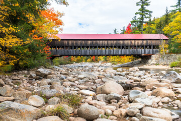 Old wooden covered bridge with a red metal roof spanning a mountain river on a partly cloudy autumn day. Beautiful fall foliage. White Mountain National Forest, NH, USA. - Powered by Adobe