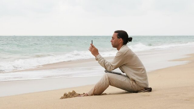 Relaxed man sit on sea beach, stare to smartphone, lean to laptop lying on knees. Suddenly water run up and catch him, notebook become wet. Hapless guy raise it up, look around, stand up and go away
