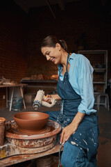 Young cheerful craft woman wearing apron working on pottery wheel with fresh wet clay in art studio