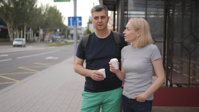 A middle-aged man and woman drink coffee from paper cups, stand at a public transport stop waiting for the bus they need.