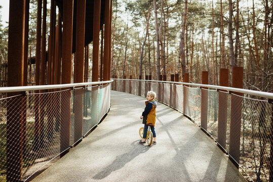 Little blond boy on a bridge in the forest with his yellow bicycle