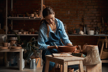 Friendly attractive female artisan wearing apron modeling ceramic tableware in pottery workshop