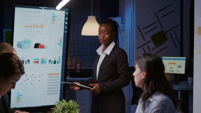 Overworked businesswoman with dark skin brainstorming financial strategy overworking in company meeting room late at night. Workaholics multi-ethnic teamwork discussing marketing deadline project