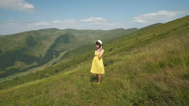 Young happy woman traveler in yellow dress standing on green grassy hillside on a windy day in summer mountains enjoying view of nature.