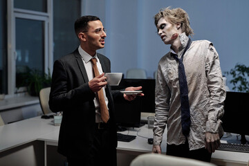 Young elegant businessman with cup of tea talking to male zombie in office